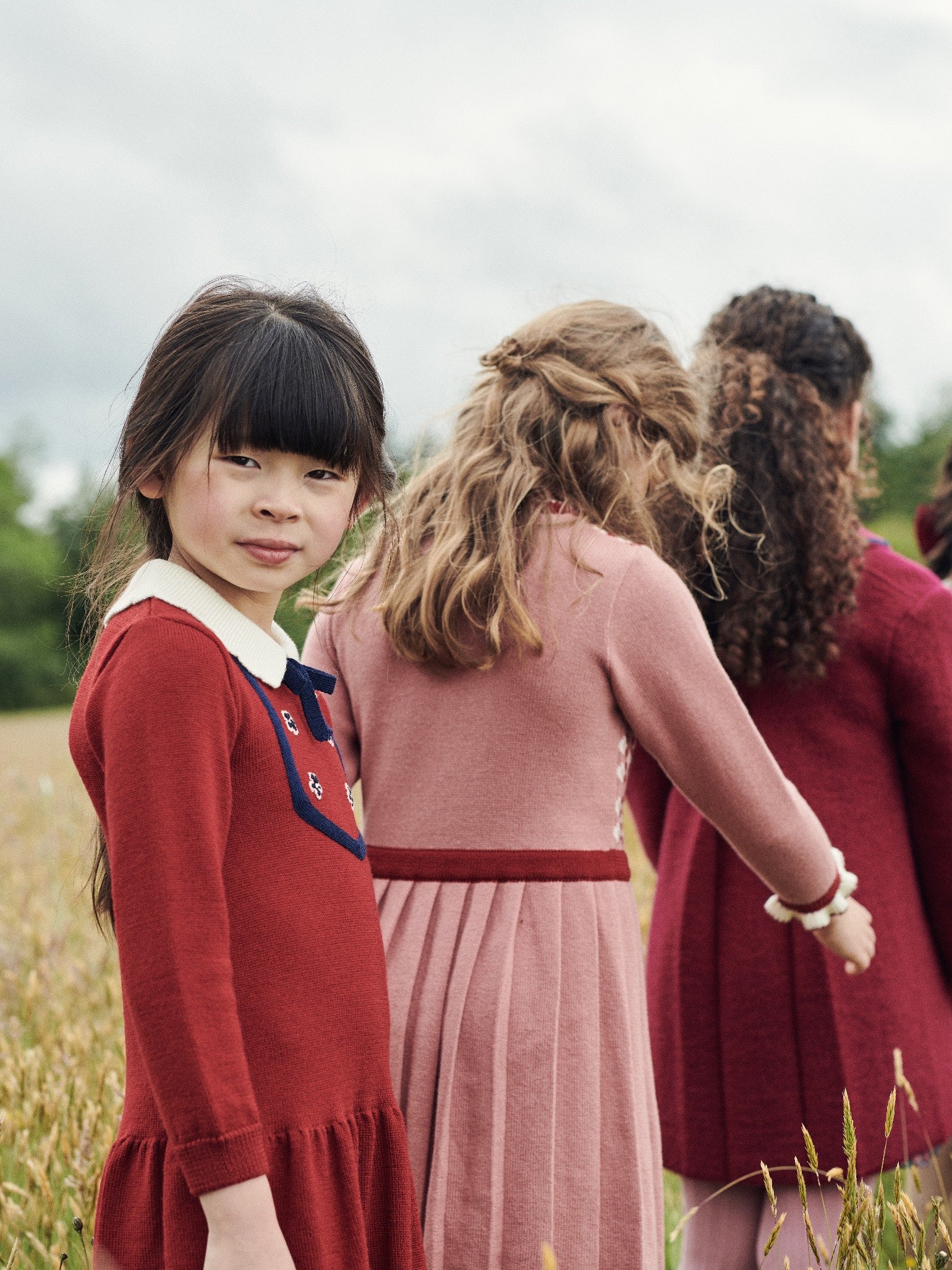 A young girl in a stylish red knit dress with navy accents stands confidently in a field, showcasing elegant children's fashion.